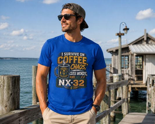 A man wearing a blue graphic t-shirt with a coffee-themed slogan stands on a wooden dock by the water. The t-shirt features a graphic with a coffee mug and the text 'I SURVIVE ON COFFEE, CHAOS, GUESS WORDS, AND NX-32'.